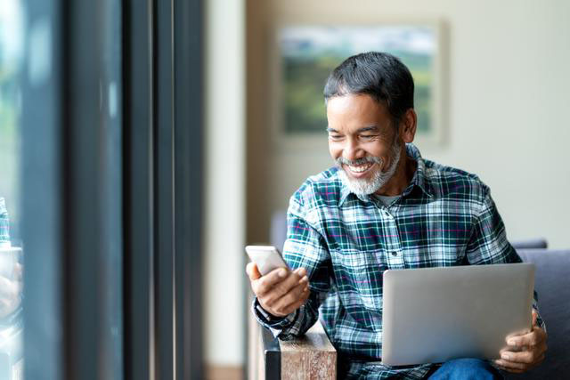 Seated man smiles at phone with laptop on lap